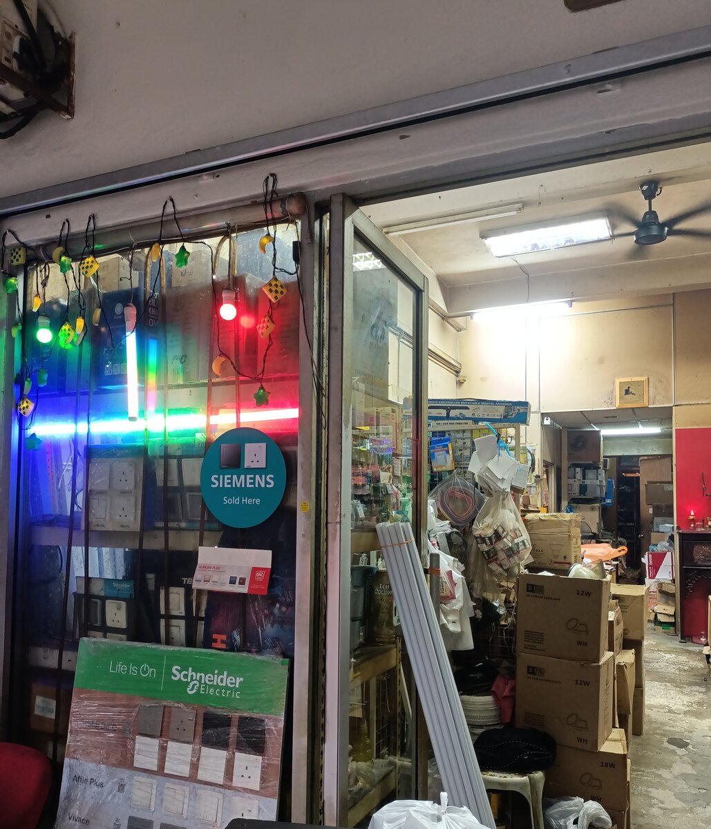 Glass shop door with coloured string lights, a round Siemens Sold Here sticker, a Schneider switch display board and stacked downlight cartons inside