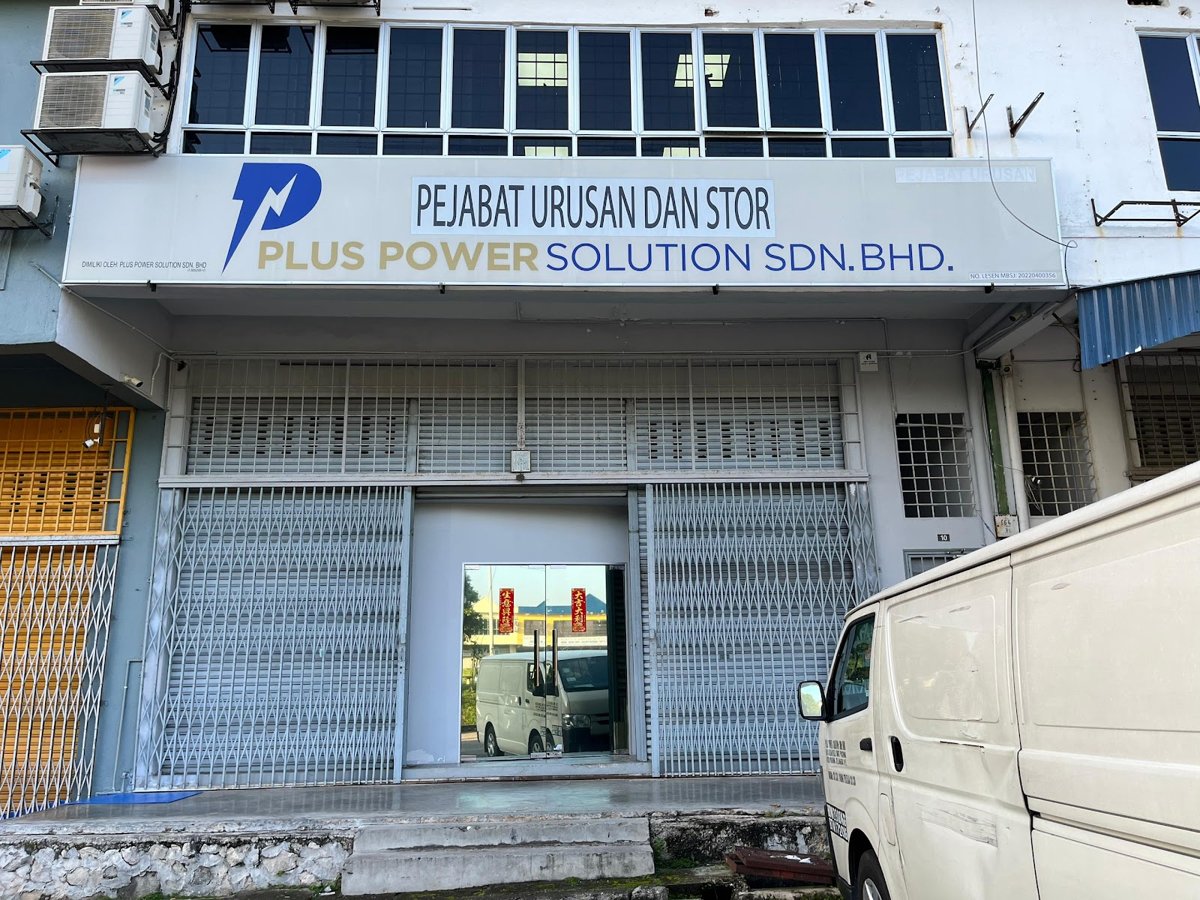 White shoplot with a Plus Power Solution Sdn Bhd signboard reading Pejabat Urusan dan Stor, with roller shutters and a glass entrance below