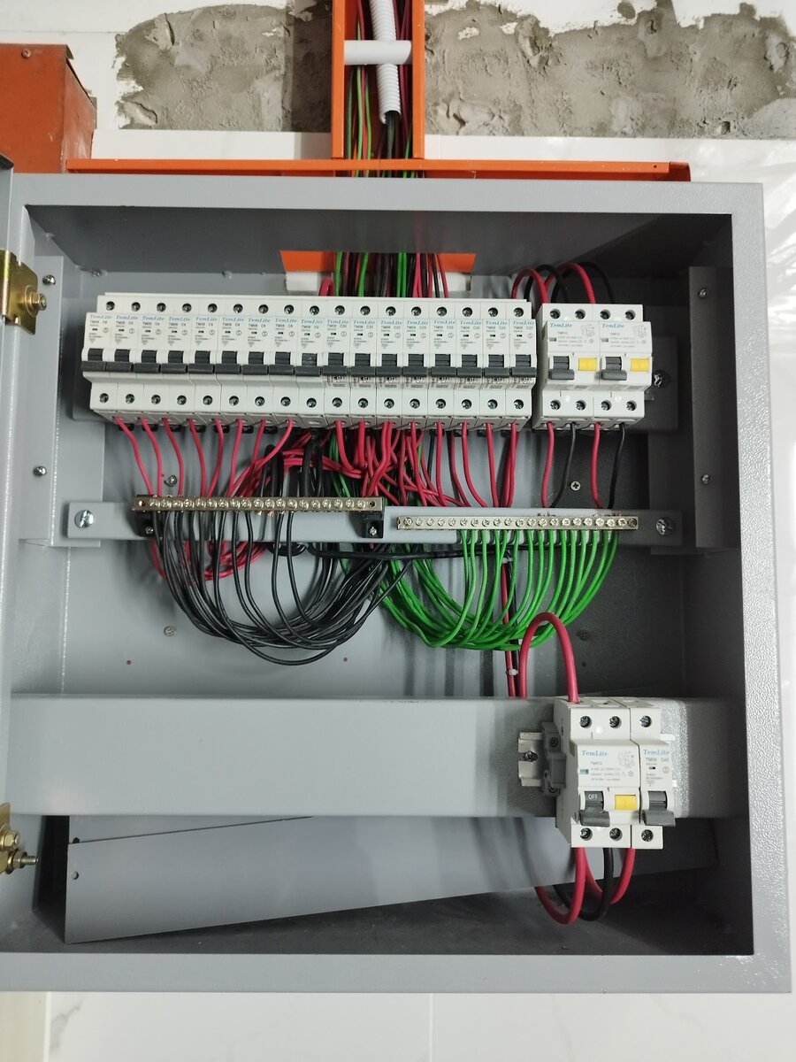 Grey metal distribution board open to show a row of miniature circuit breakers, RCCBs with yellow test buttons and neatly dressed red, black and green cables