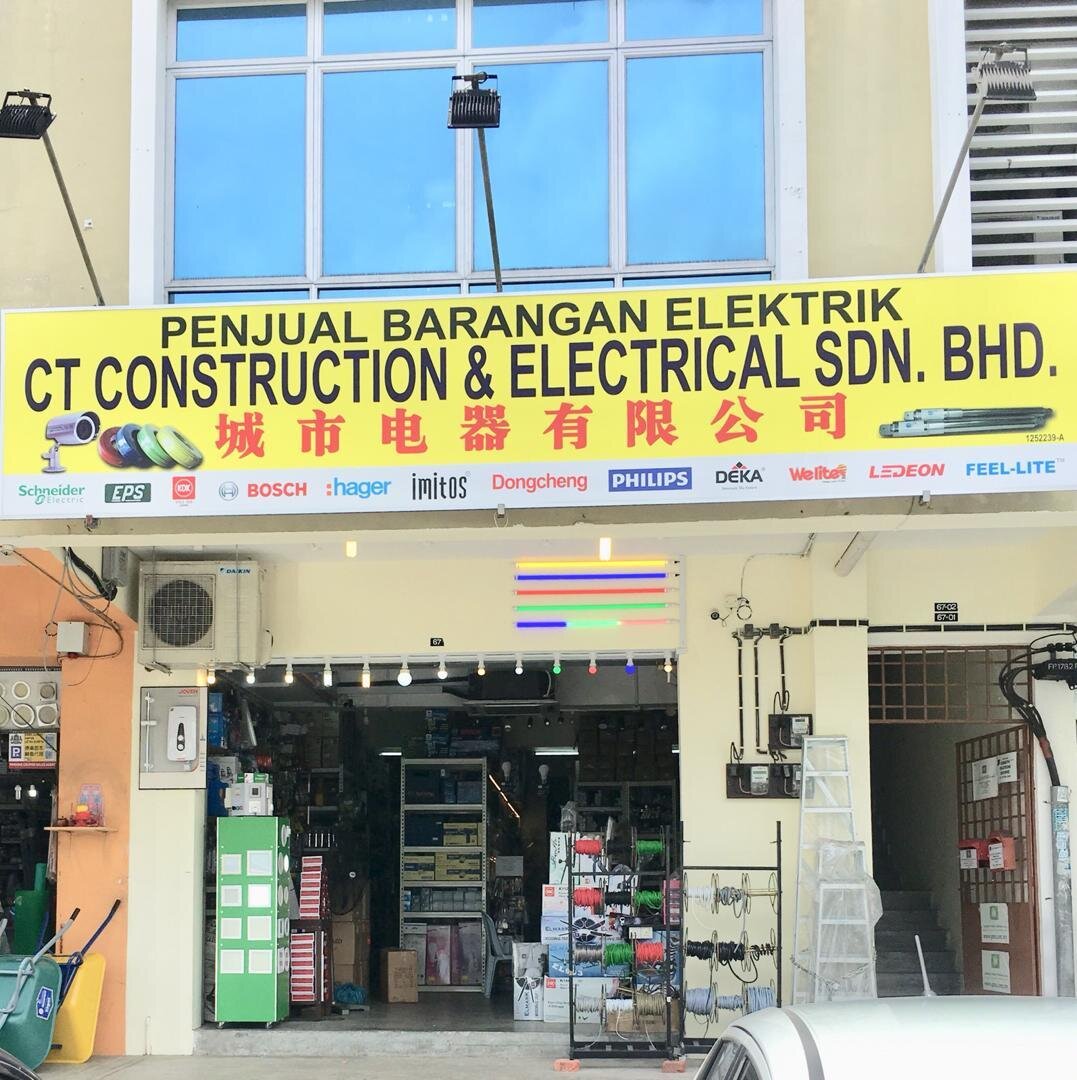Yellow signboard reading Penjual Barangan Elektrik CT Construction & Electrical Sdn Bhd above an open electrical shop, with a row of brand logos including Schneider Electric, Hager, Philips and Deka, and racks of cable reels outside