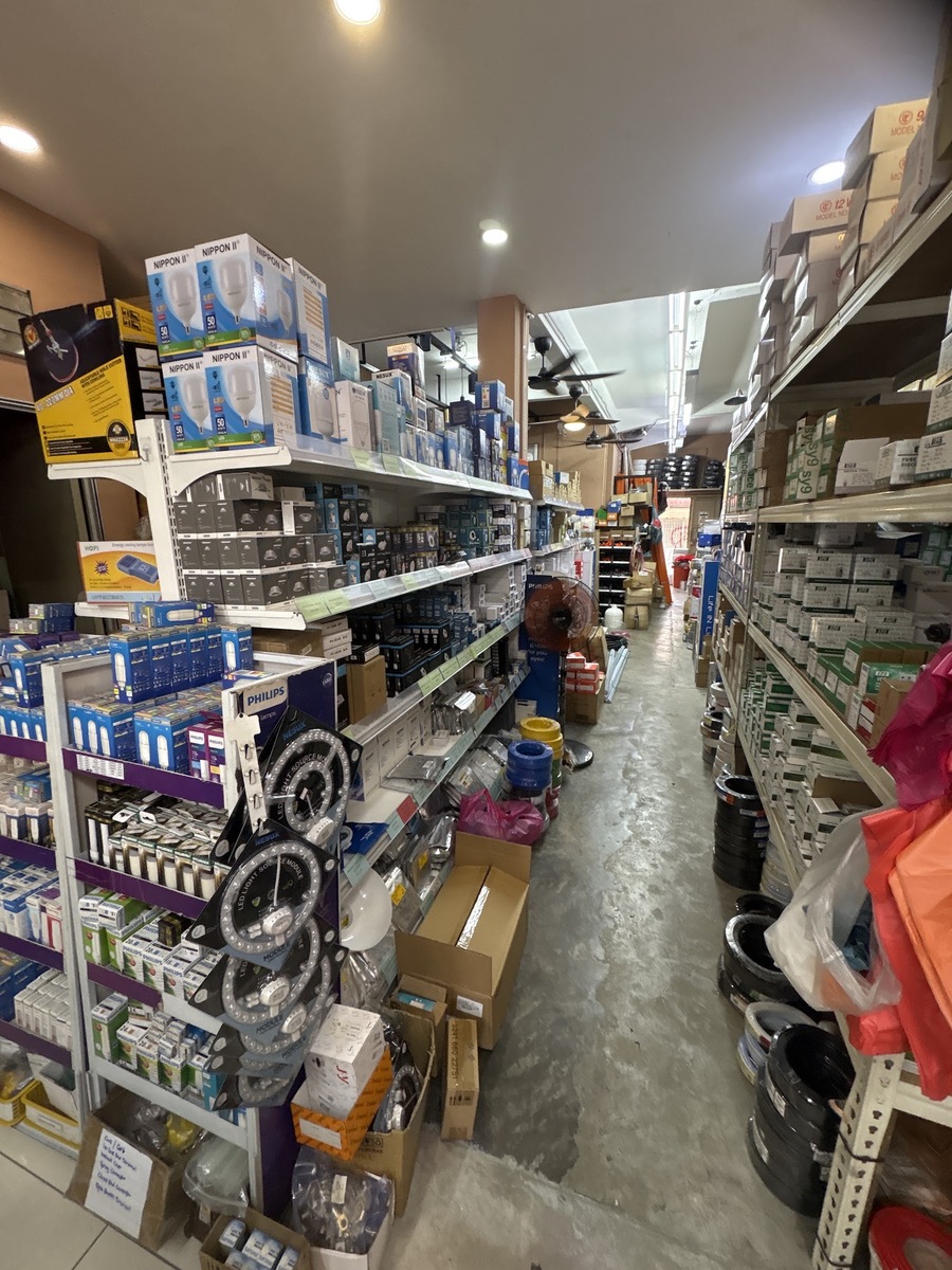 Narrow shop aisle with shelves of boxed LED bulbs and wiring accessories on both sides, a Philips display stand and coils of cable on the floor