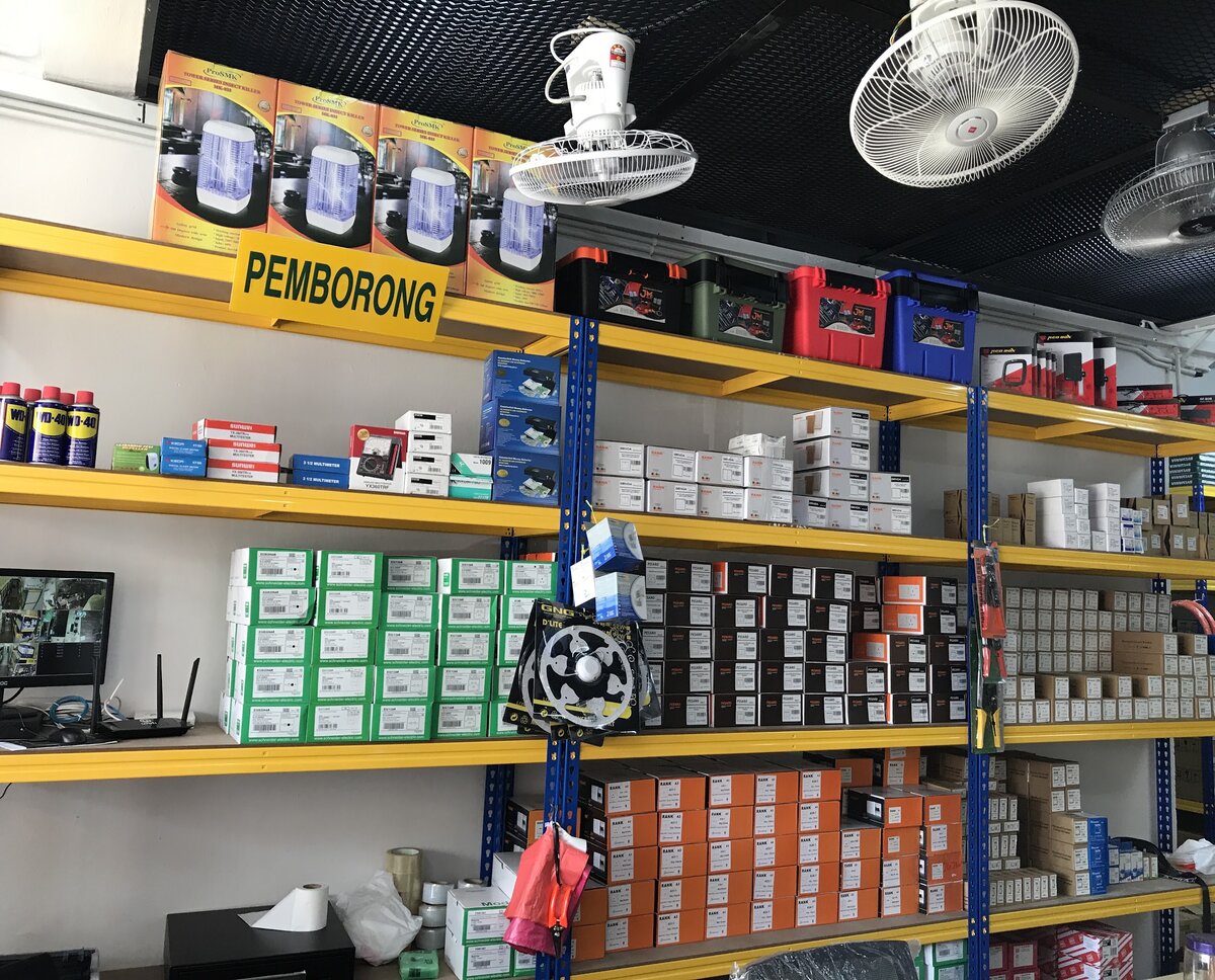 Yellow and blue steel shelving stacked with green Schneider boxes, black and orange Rank boxes and red-and-white multimeter boxes, a yellow Pemborong sign on the top shelf and fans mounted on the ceiling