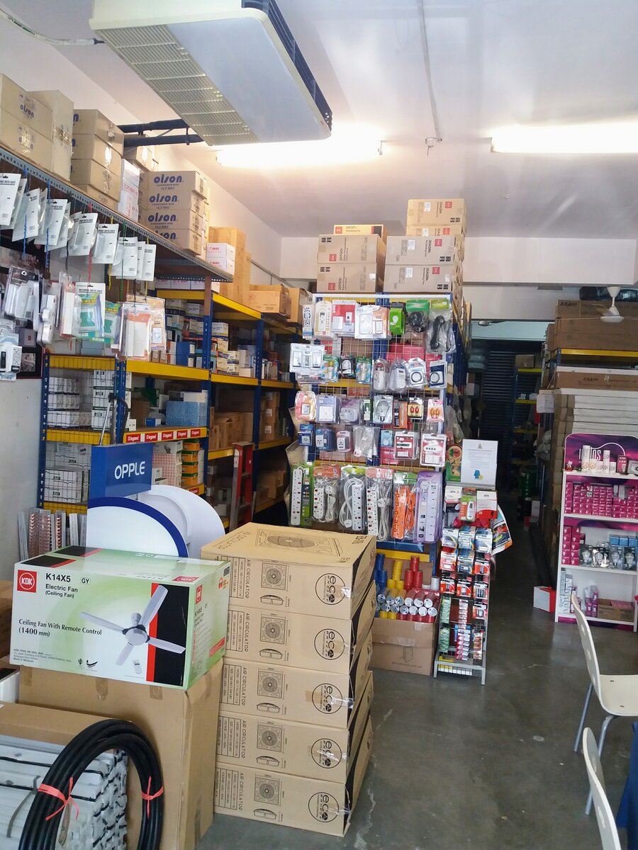 Shop interior with shelves of boxed wiring accessories under stacked Olson consumer box cartons, a wire rack of extension sockets, a blue Opple sign, a KDK K14X5 ceiling-fan carton and a stack of air-circulator boxes