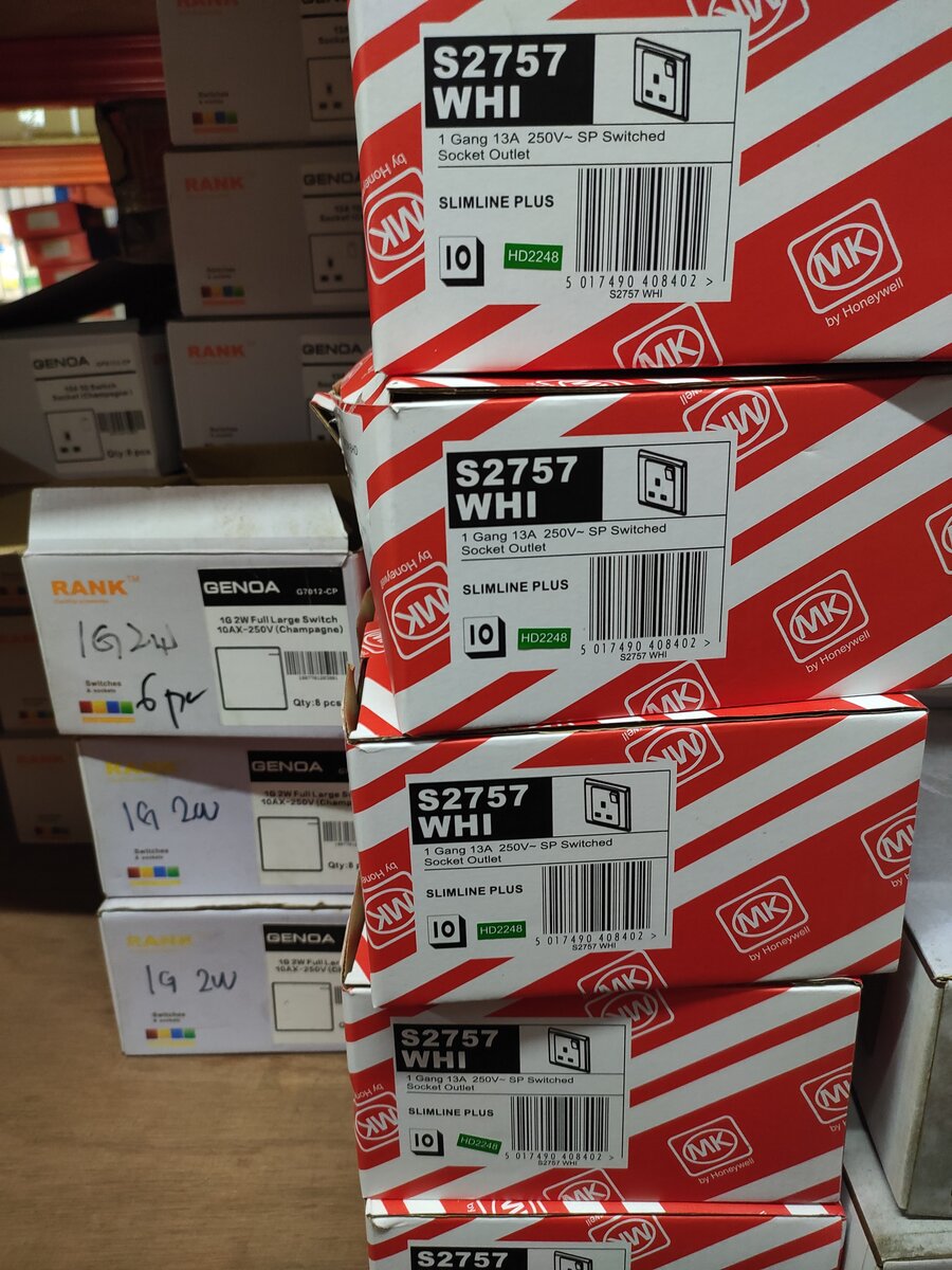 Stack of red and white MK boxes labelled S2757 WHI 1 Gang 13A switched socket outlet, beside white Rank Genoa switch boxes with handwritten 1G 2W labels