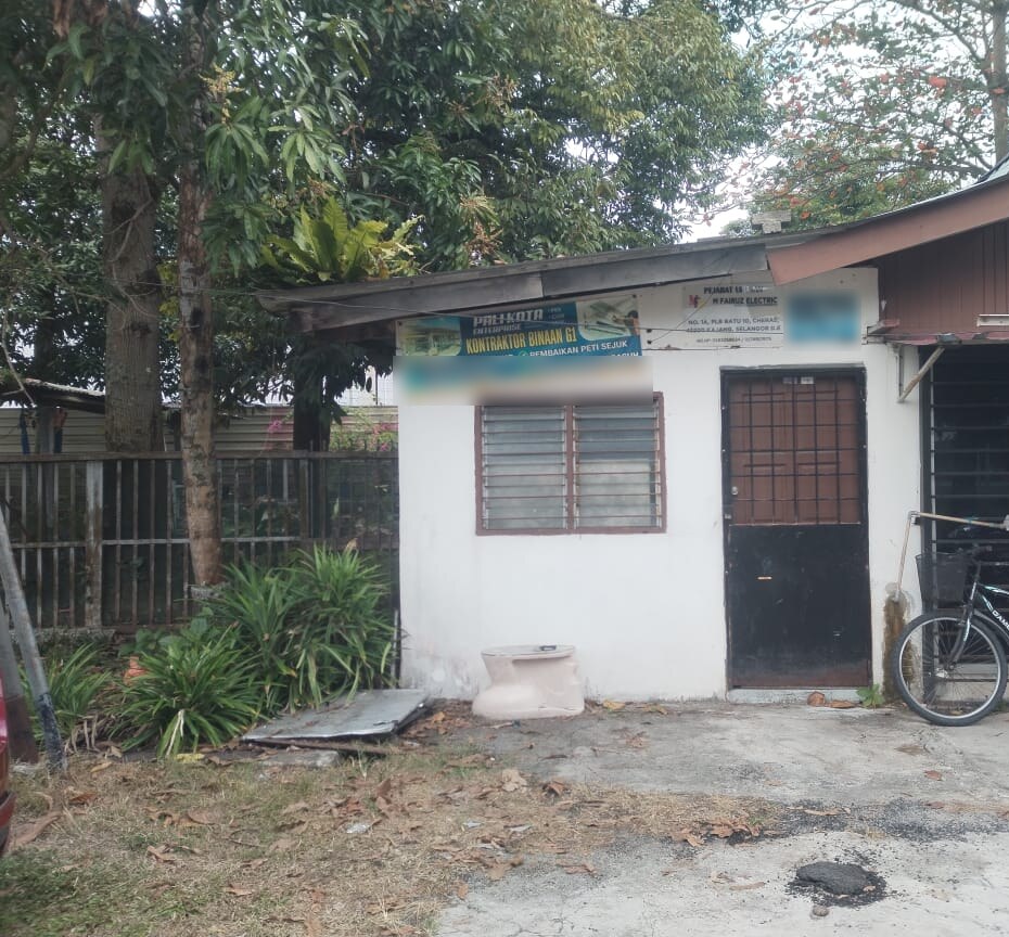 Small white single-storey hut with a barred window and a dark door, a large blue banner for an aircond and appliance repair business and a smaller white M Fairuz Electric sign above the door, trees behind