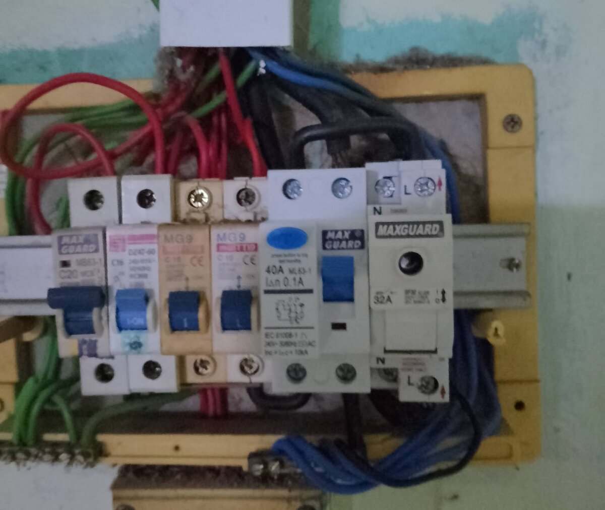 Close view of an old yellow-framed distribution board on a green wall, with four small MCBs of mixed brands, a white Maxguard RCCB with a blue test button and a Maxguard 32A device with an indicator lamp
