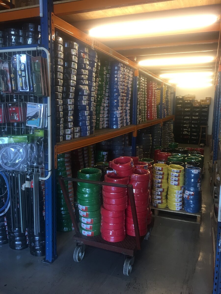 Storage aisle with shelves of cable coils in red, green, yellow and blue, a trolley of red cable, and conduit and tools on mesh racks