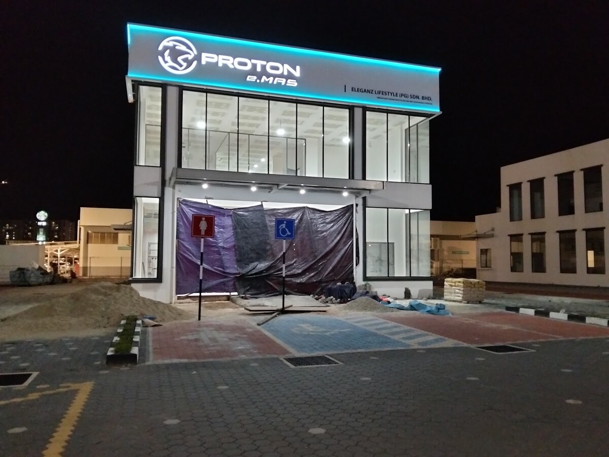 Two-storey glass-fronted Proton showroom lit at night with its entrance under tarpaulin and paving work in front