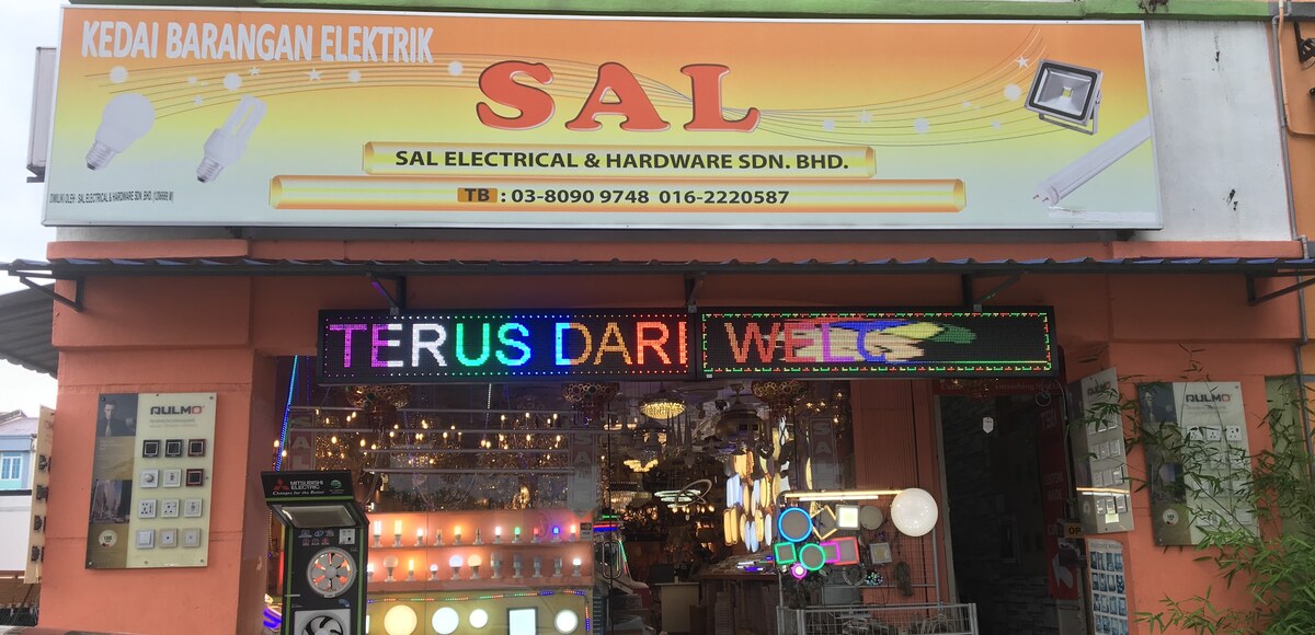 Yellow SAL signboard above a scrolling LED sign reading Terus Dari, a lit shop window of bulbs and panel lights, and switch display boards on both sides