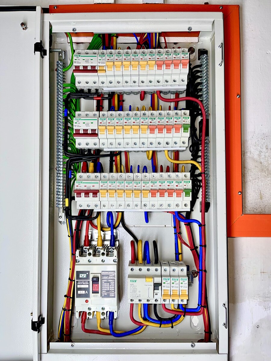 Open grey distribution board with three rows of EPS miniature circuit breakers, each row starting with a dark-red three-pole breaker followed by yellow and salmon-pink handles, above a moulded-case main breaker and an RCCB, with neatly dressed coloured cables