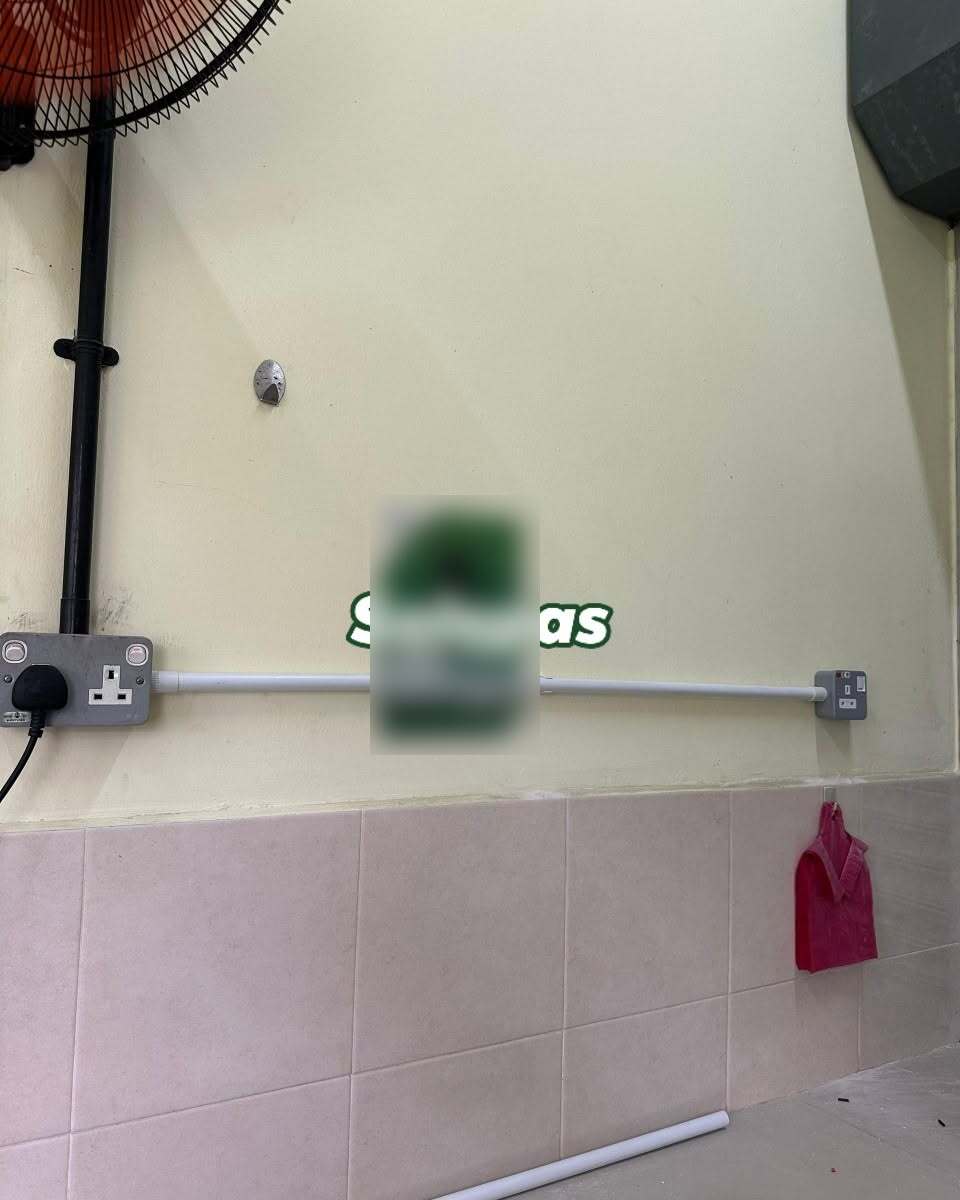 Cream wall above pink tiles with a grey metal-clad socket with a plug in it on the left, a white conduit running right to a small new grey socket, a wall fan at the top left and a blurred notice in the middle