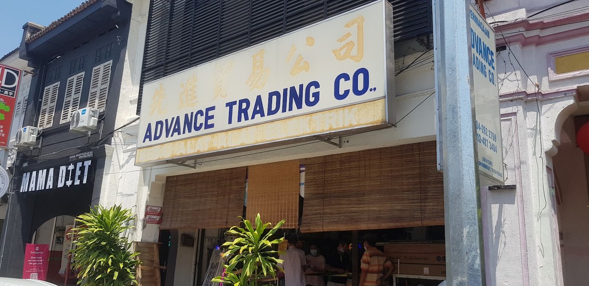 Shophouse with a cream signboard reading Advance Trading Co. in blue and Chinese characters, bamboo blinds over the entrance and people at the entrance