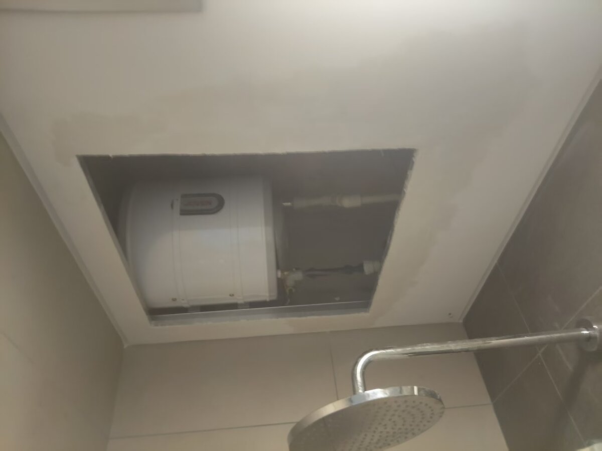Looking up through an open square hatch in a bathroom ceiling at a white cylindrical water heater, with a rain shower head on the wall below