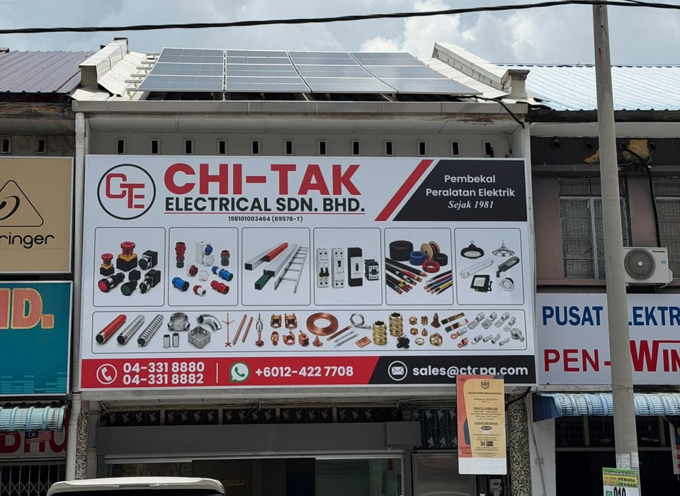 Shophouse front with a white "CHI-TAK ELECTRICAL SDN. BHD." signboard showing pictures of switches, cable trunking, circuit breakers, cables, lights and fittings, phone numbers on a red strip, and solar panels on the roof