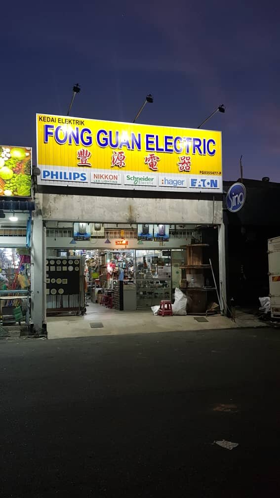 Shop at dusk with a yellow 'Kedai Elektrik Fong Guan Electric 豐源電器' signboard lit by floodlights, a row of brand logos underneath and an open shopfront showing light fittings