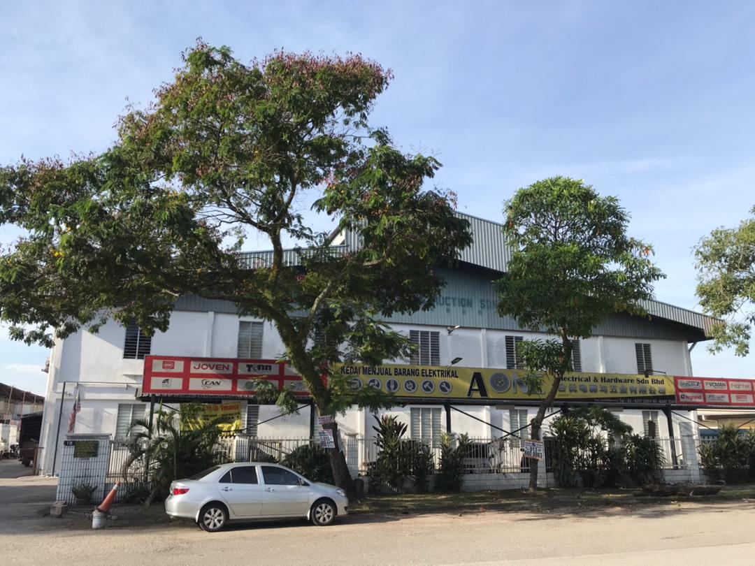 White single-storey warehouse with a grey roof behind trees, with a long yellow banner reading A ONE Electrical & Hardware Sdn Bhd and red boards of brand logos along the fence line
