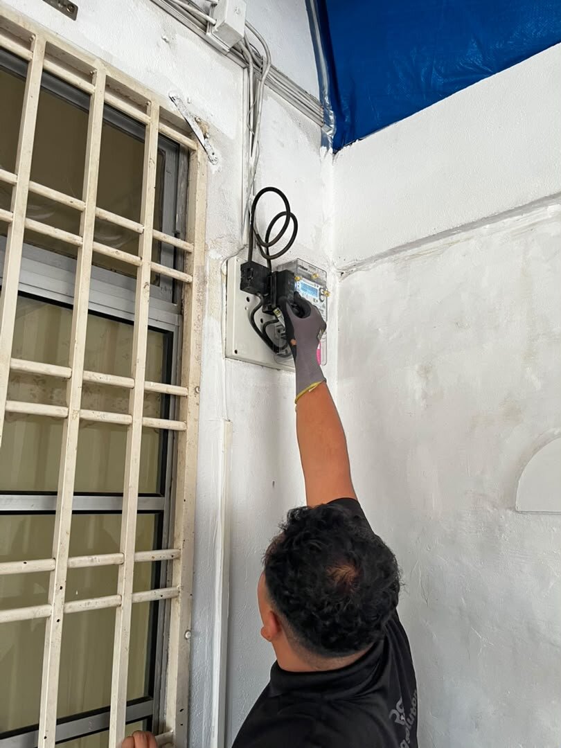 Technician reaching up to an electricity meter mounted on an outside wall beside a window grille