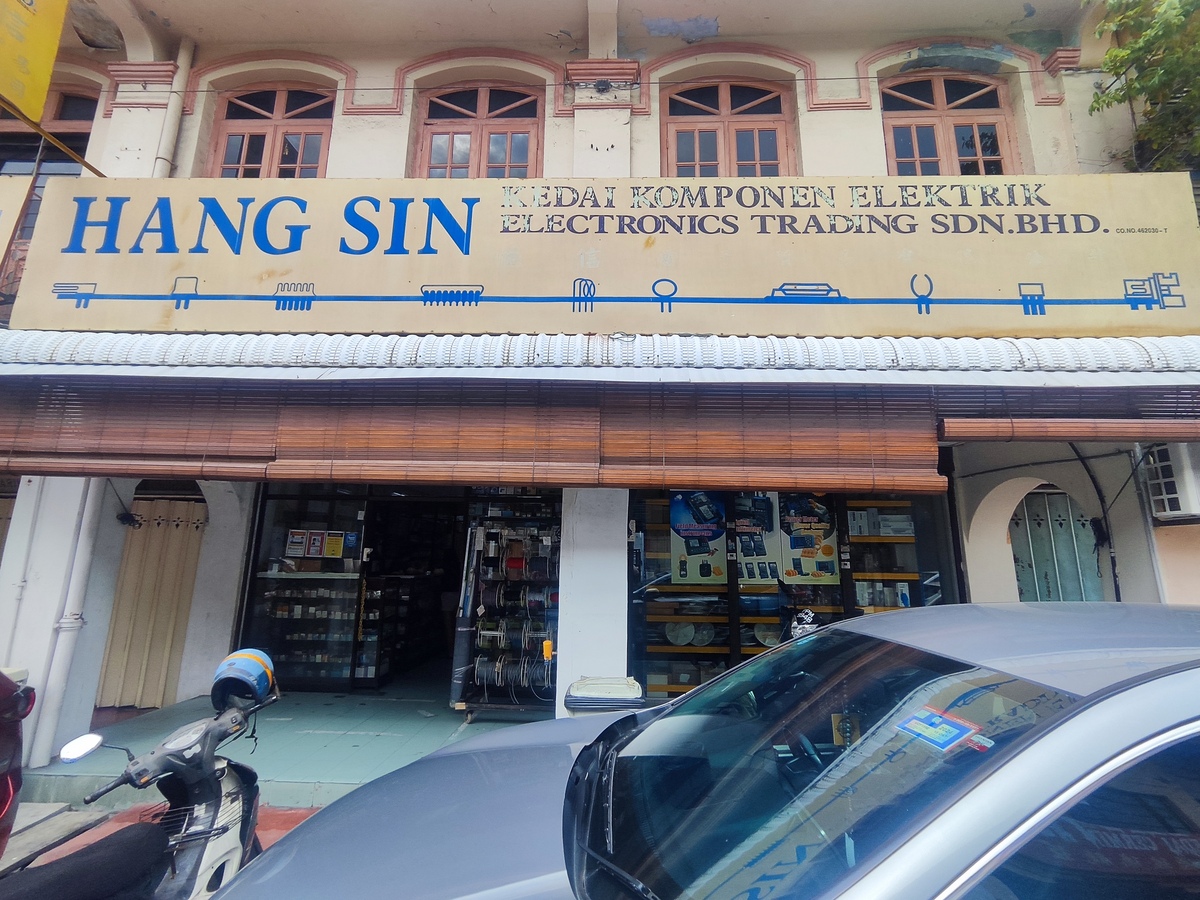 Old shophouse with a long cream signboard reading HANG SIN, Kedai Komponen Elektrik, Electronics Trading Sdn Bhd, decorated with electronic component symbols, above an open shopfront with wire reels and display shelves