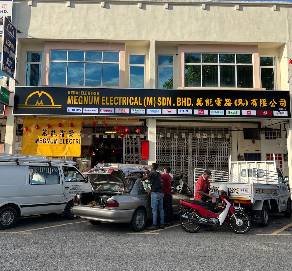 Black Megnum Electrical signboard with a row of brand logos above two shop units, with a van and a pickup parked in front
