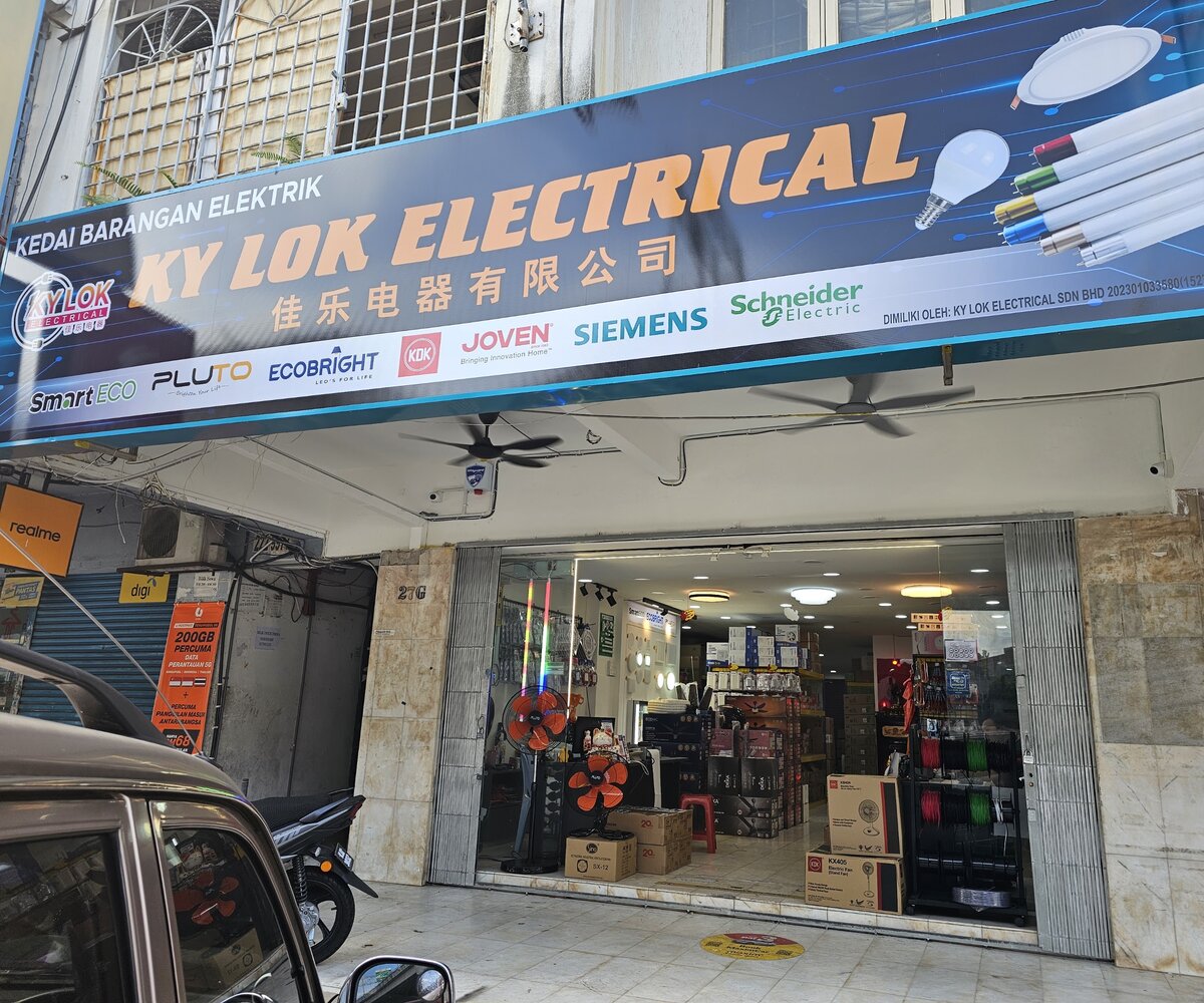 Blue signboard reading Kedai Barangan Elektrik, Ky Lok Electrical and 佳乐电器有限公司 with brand logos, over an open shopfront with fans and a rack of wire reels