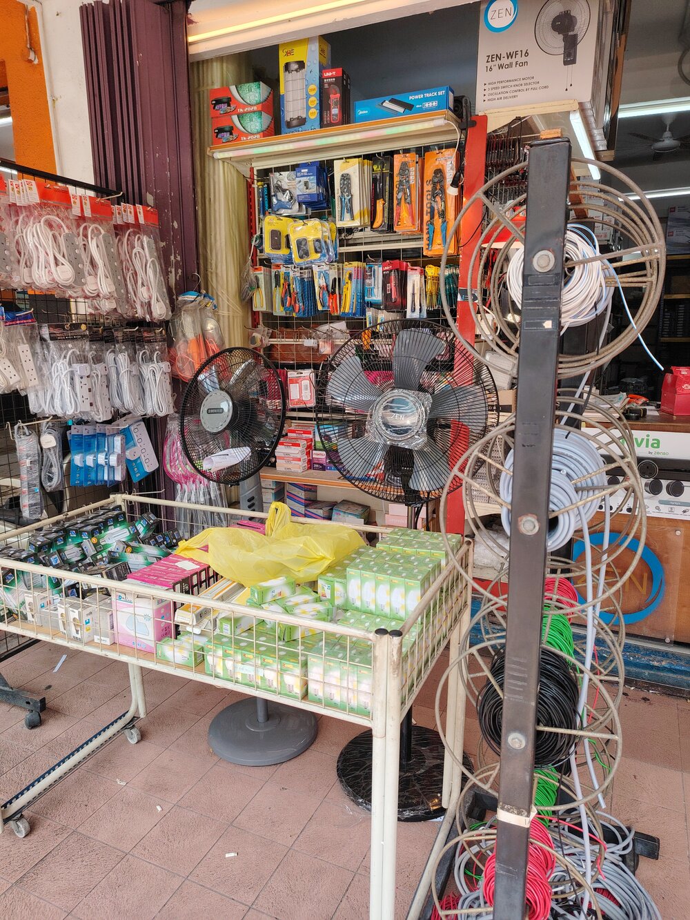 Reels of white, grey, red, green and black cable on a stand beside two stand fans (Zen and SiberAir), a wall of hanging tools and extension sockets, and a wire bin of boxed bulbs