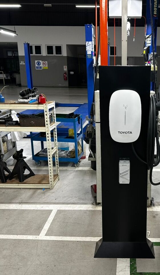 Black pedestal with a white Toyota wall charger in a workshop bay, with shelves, jack stands and vehicle lifts behind