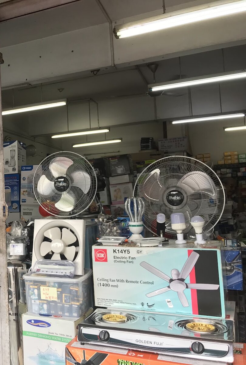 Shop entrance with two stand fans, an exhaust fan, LED bulbs and a boxed KDK ceiling fan stacked on boxes, and shelves of electrical stock behind