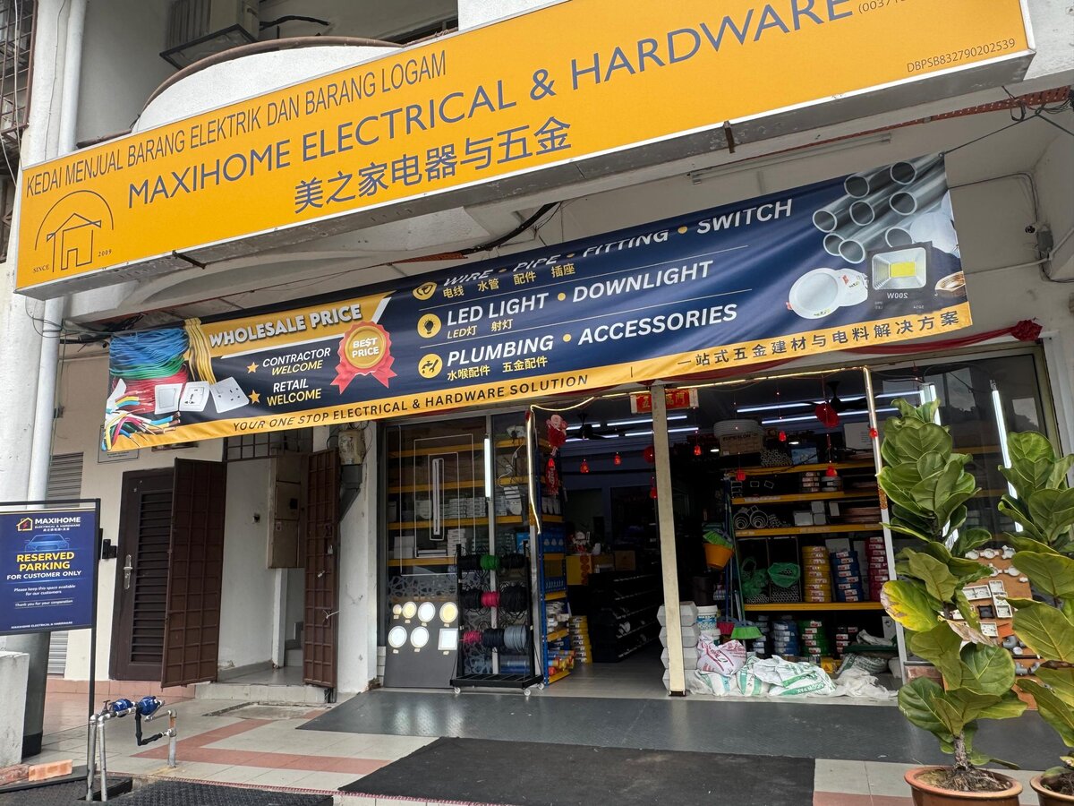 Shopfront with a yellow Maxihome Electrical & Hardware signboard, a dark blue banner listing wire, pipe, fitting, switch, LED light, downlight and plumbing, and racks of goods inside