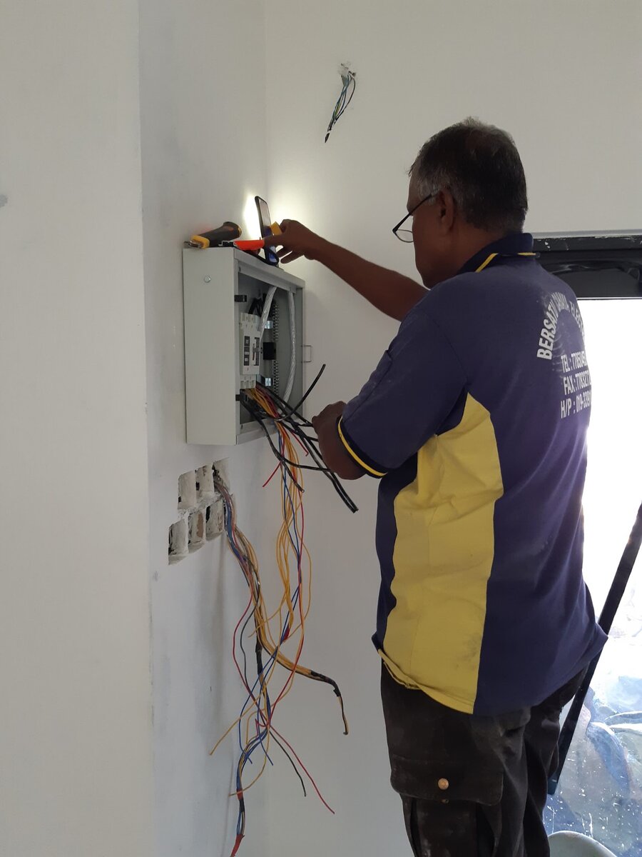 Technician seen from behind and to the side, wearing a navy and yellow shirt printed with Bersatu Cahaya Electric, working on a surface-mounted distribution board with loose coloured wires and socket boxes below