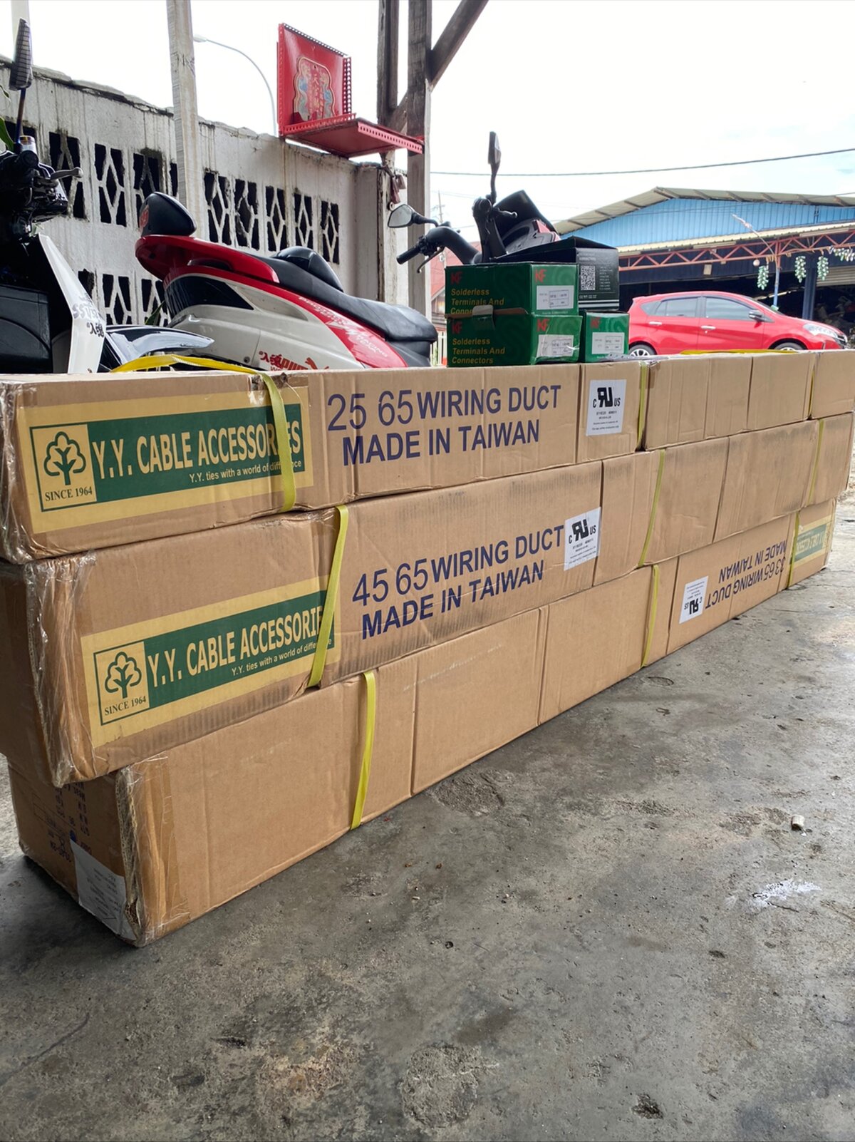 Stacked brown cartons printed 'Y.Y. Cable Accessories' and '25 65 Wiring Duct, Made in Taiwan', with green boxes of KF solderless terminals on top and parked motorcycles behind