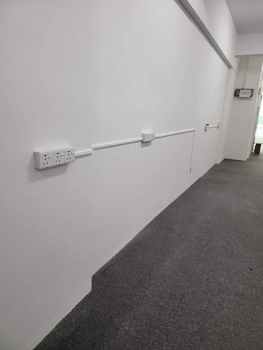 Long carpeted office corridor with twin wall sockets joined by white surface trunking along the wall