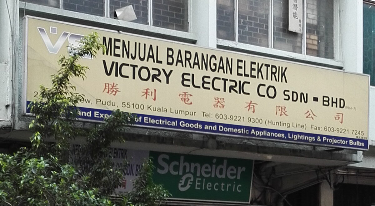 Faded cream shop signboard reading Menjual Barangan Elektrik, Victory Electric Co Sdn Bhd and Chinese characters, with the address and phone numbers along its lower edge and a blue strip describing its trade, above a green Schneider Electric sign, partly behind a small tree