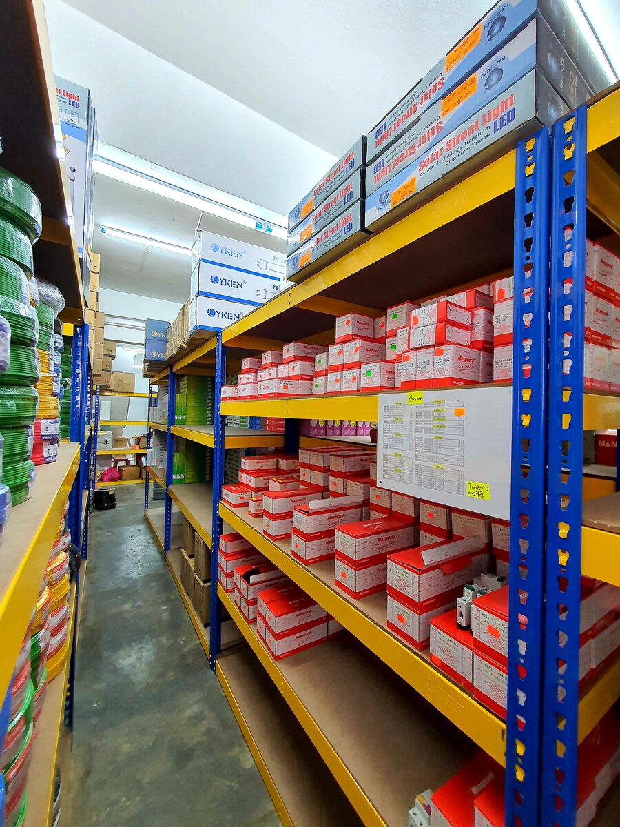 Yellow and blue steel shelving stacked with red and white Maxguard boxes, a printed price sheet, and green cable coils along an aisle