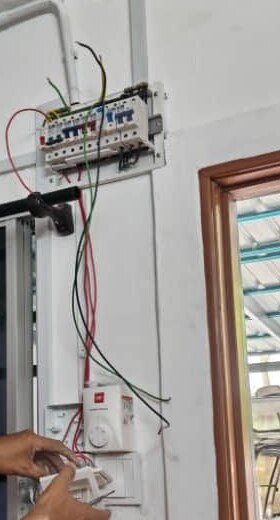 White wall with a small open distribution board holding a row of breakers, loose cables hanging below it to a wall switch and a round controller, a window frame to the right and a hand at the bottom