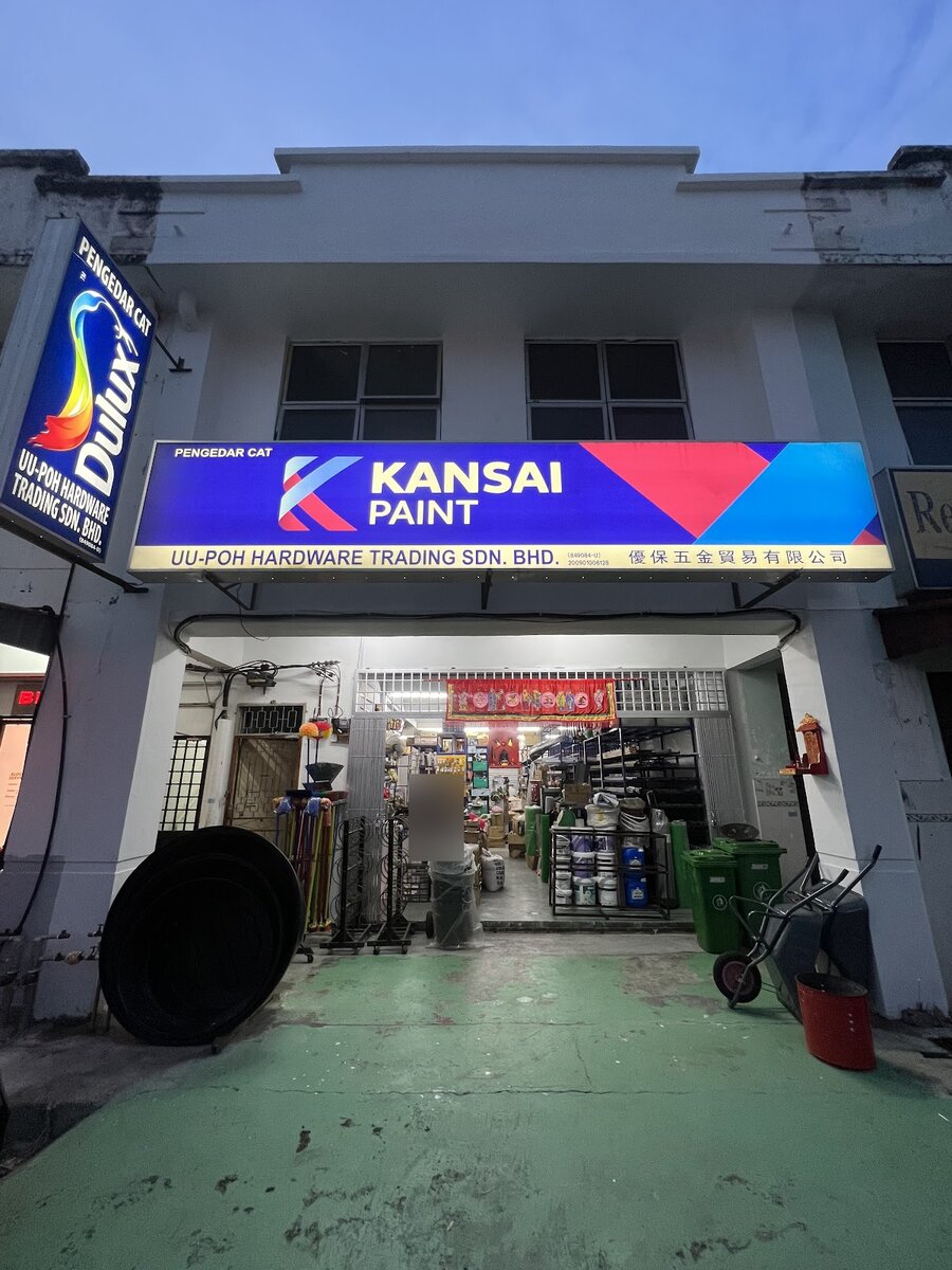 Two-storey shophouse with a lit Kansai Paint signboard reading UU-Poh Hardware Trading Sdn. Bhd. and a Dulux sign, paint tins and wheelbarrows at the entrance