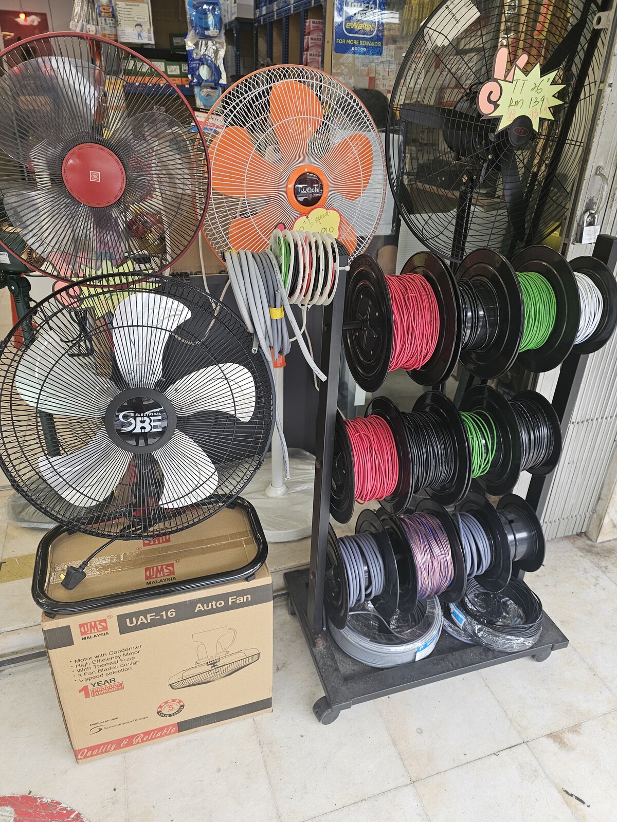 Stand and floor fans beside a black rack holding reels of red, black, green, white, grey and purple wire, with a UMS auto fan carton