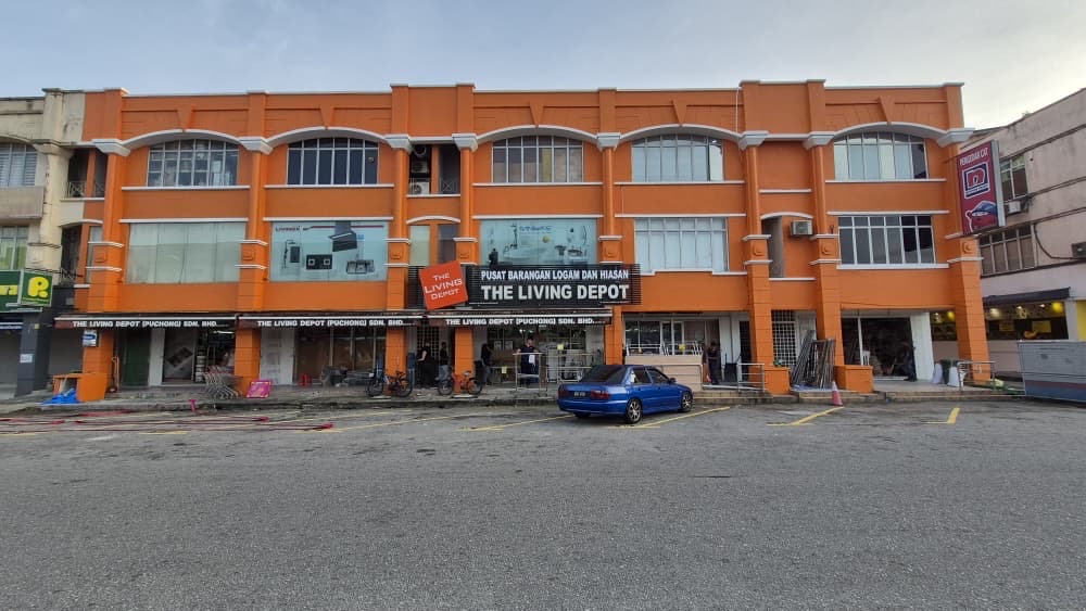 Orange three-storey shophouse block with a sign reading Pusat Barangan Logam dan Hiasan, The Living Depot, above a row of shopfronts and a car park