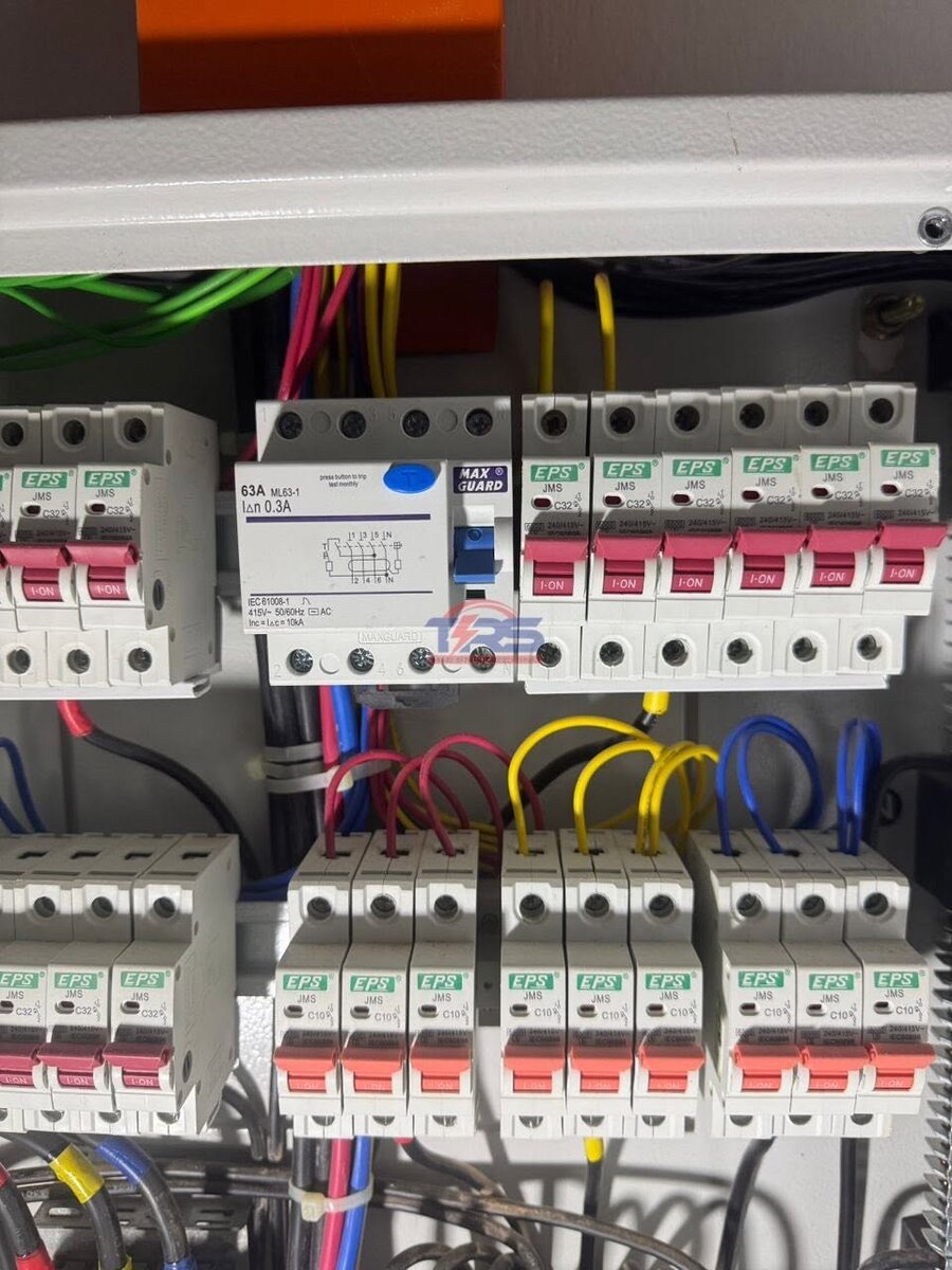 Open metal distribution board with a white four-pole Maxguard RCCB rated 63A 0.3A next to green-labelled EPS MCBs, rows of MCBs below and red, yellow and blue cables looping between them