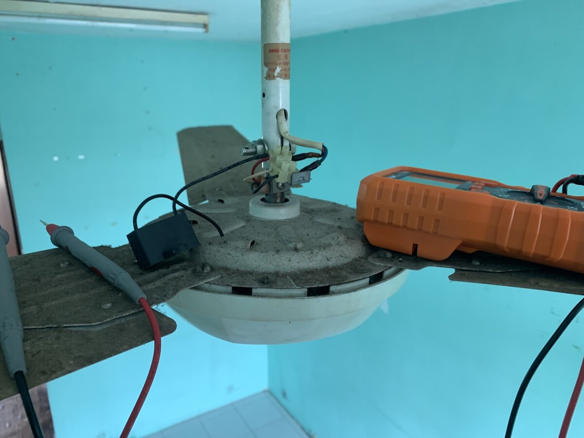 Top of a ceiling fan motor on its downrod, with its blades attached, test leads clipped to the terminal block and an orange multimeter resting on a blade, against a turquoise wall