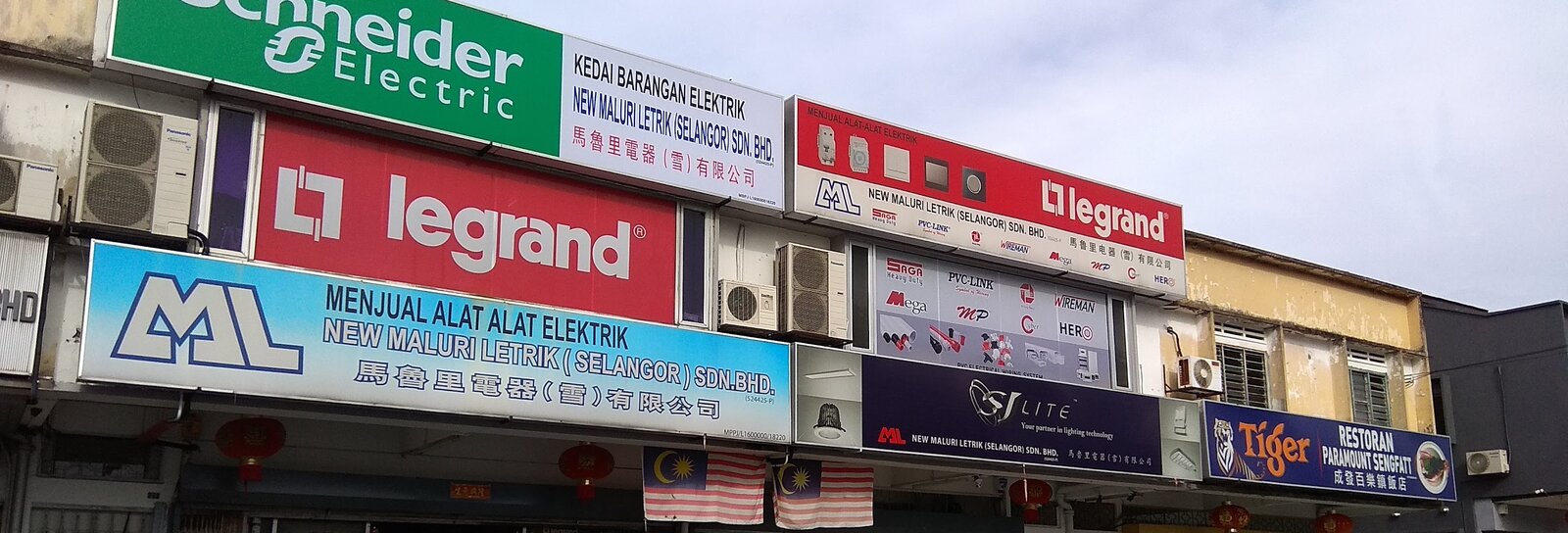 Row of shop signboards: green Schneider Electric, red Legrand, blue ML New Maluri Letrik, a panel of PVC conduit brand logos and a navy SJ Lite sign