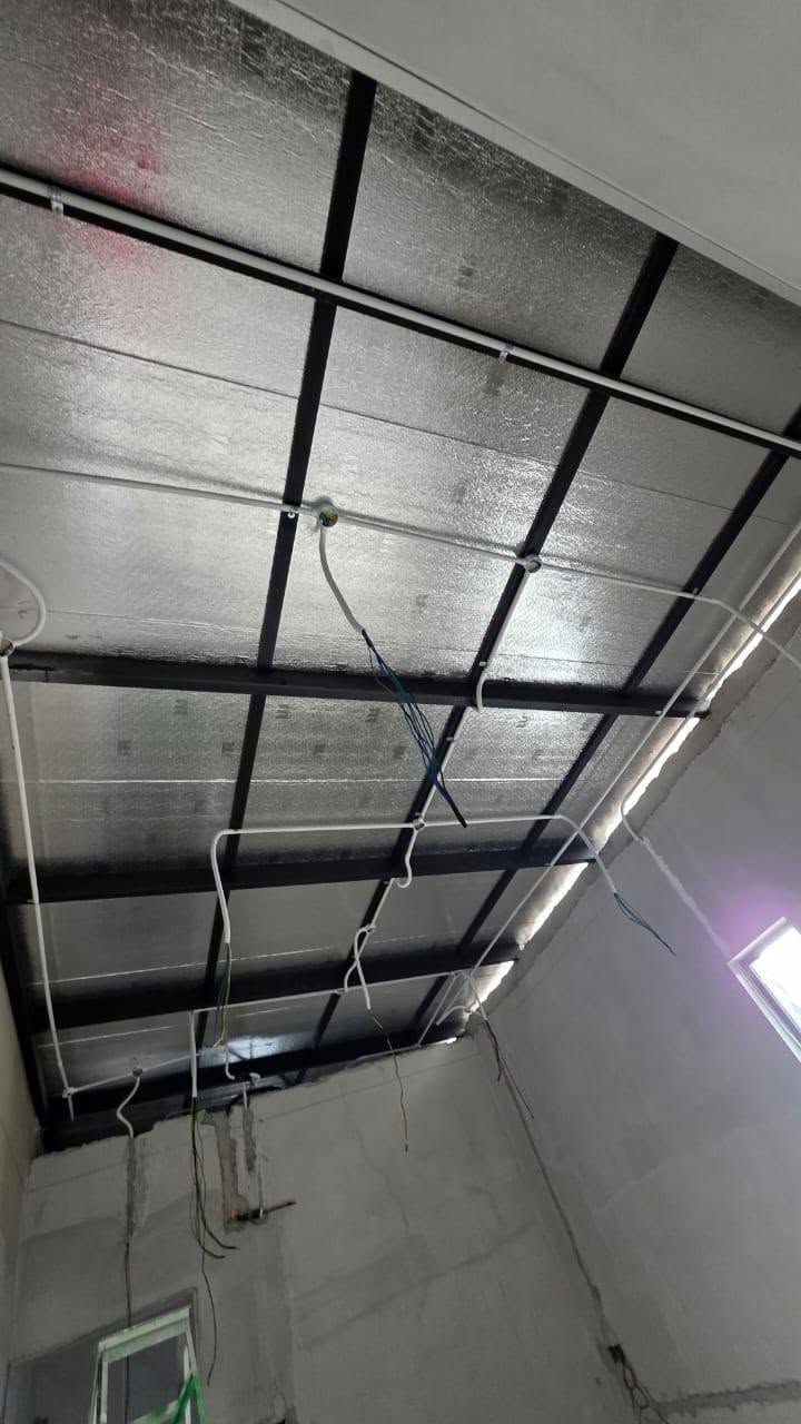 Looking up at a roof space with foil insulation, black metal ceiling framing and white conduit and cables clipped along it