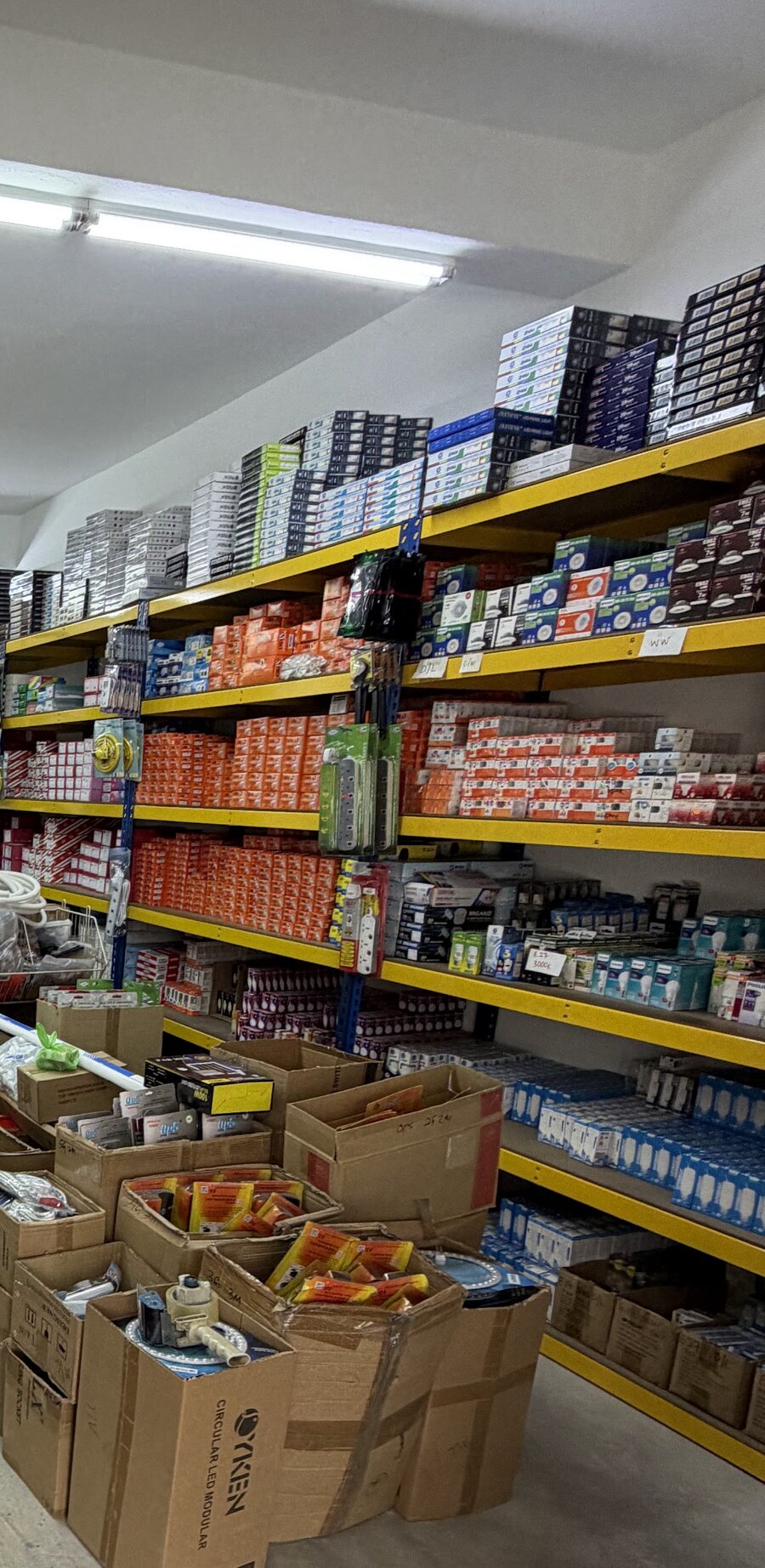 Yellow steel shelves stacked to the ceiling with boxes of LED panel lights, downlights, bulbs and orange boxed accessories, with extension sockets hanging and open cartons on the floor