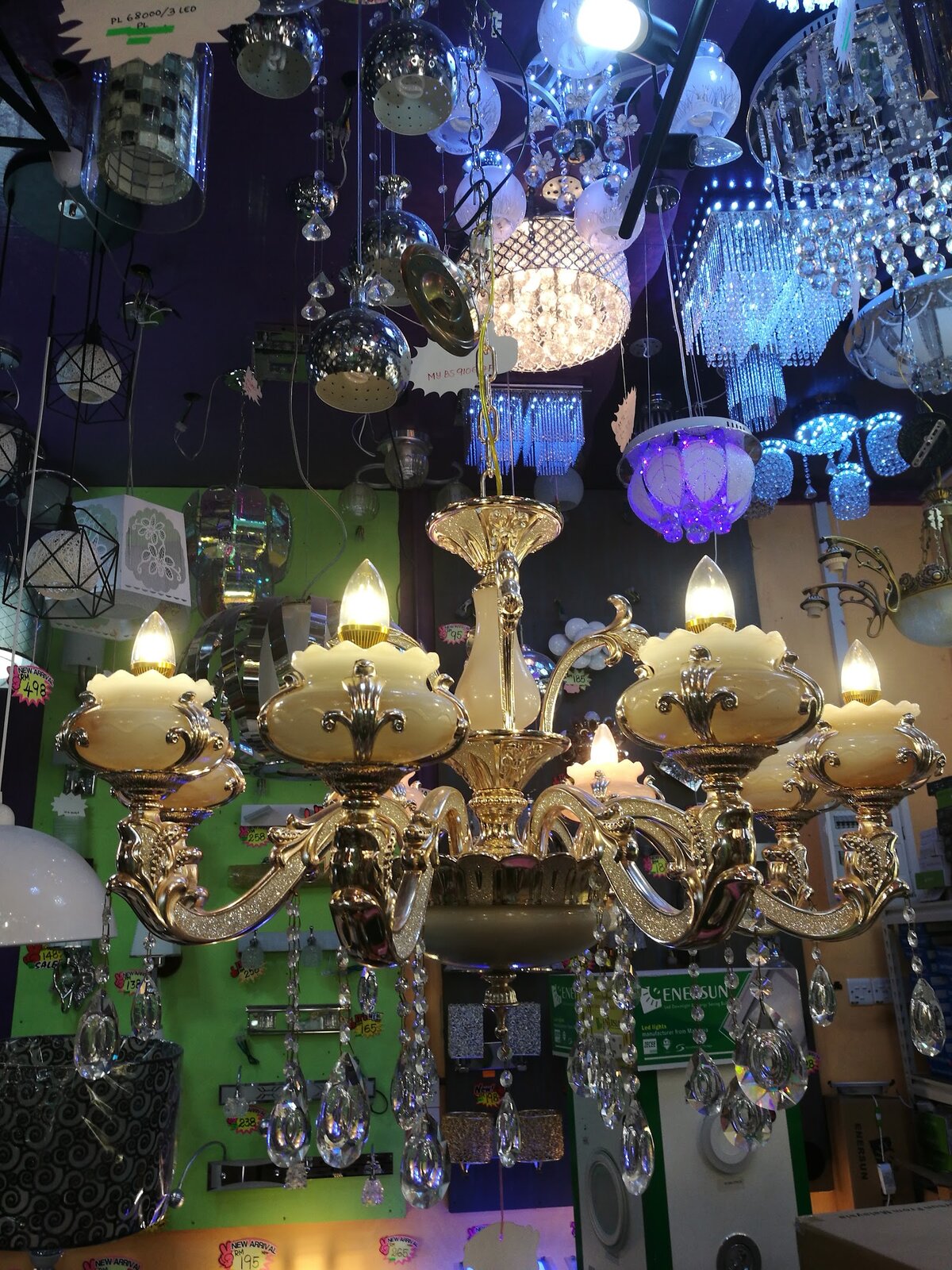 Gold chandelier with candle bulbs in front of a wall of pendant lamps, crystal chandeliers and wall lights