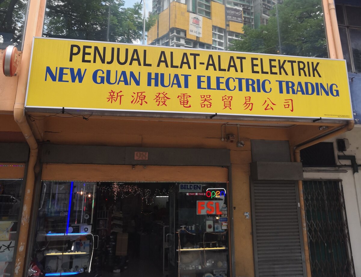 Yellow signboard reading Penjual Alat-Alat Elektrik New Guan Huat Electric Trading with red Chinese characters, above a glass shopfront with lit display windows and brand stickers on the door