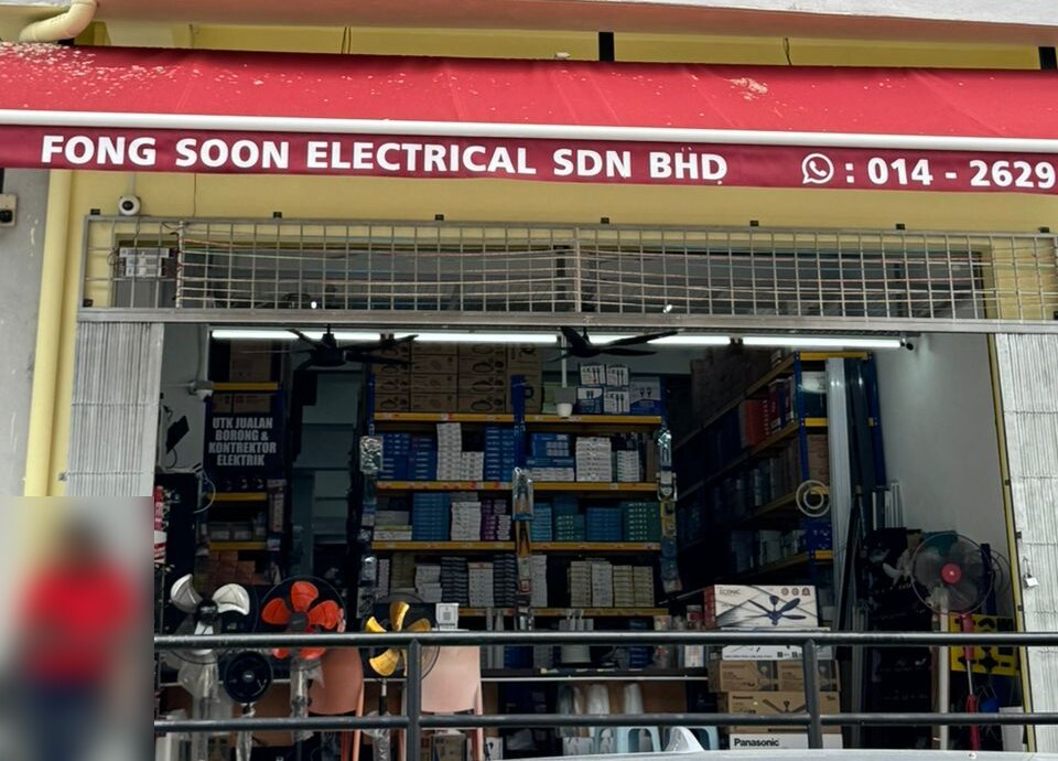 Shop with a red awning reading Fong Soon Electrical Sdn Bhd, open shutter, shelves of boxed goods, ceiling fans and stand fans inside