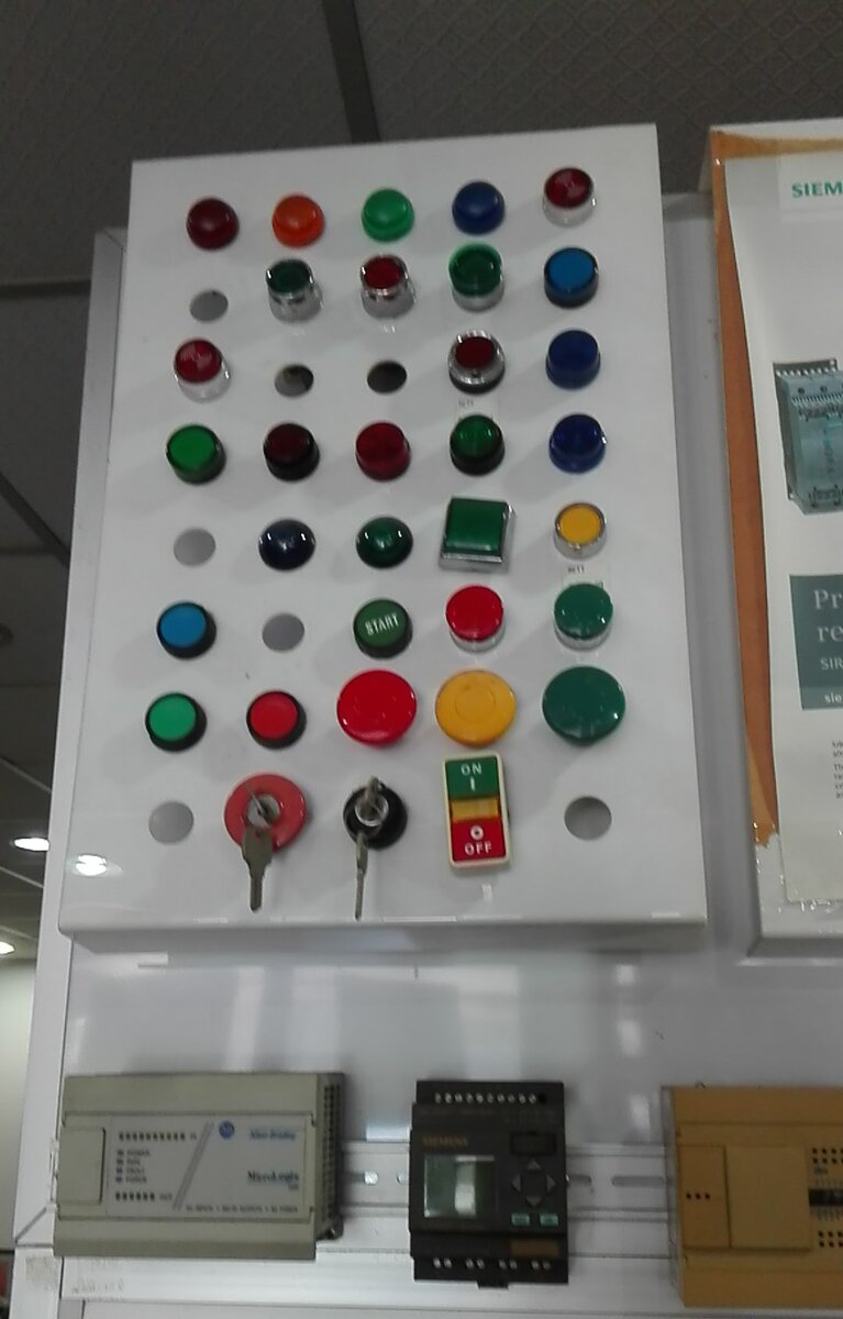 White panel fitted with coloured pilot lamps, push buttons, mushroom stop buttons and key switches, with three small PLC modules on a DIN rail below
