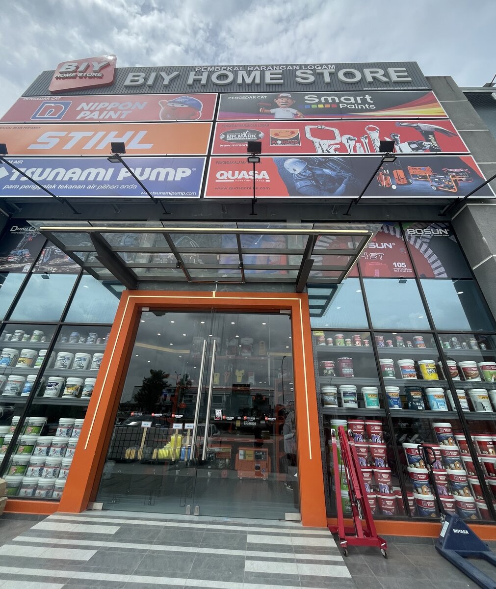 Glass shopfront with an orange frame under a "BIY Home Store" fascia and brand signboards, shelves of paint tins behind the windows