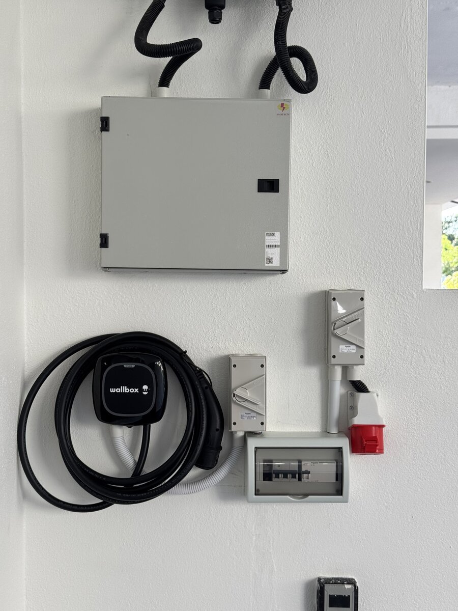 Black Wallbox wall charger with a coiled Type 2 cable on a white wall, beside two grey rotary isolators, a small enclosed breaker board and a red industrial socket, under a grey metal distribution board
