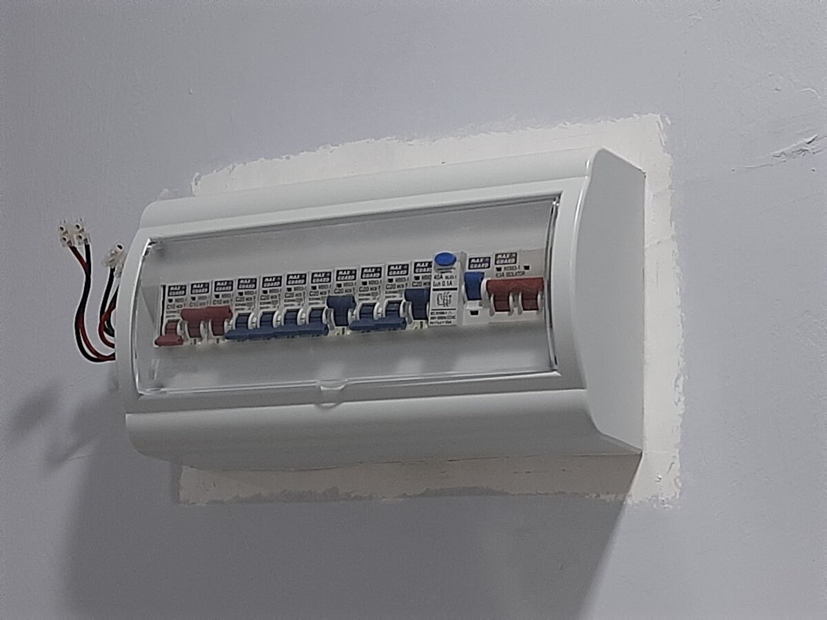 White surface-mounted distribution board with a row of circuit breakers and two cable ends on a freshly plastered wall