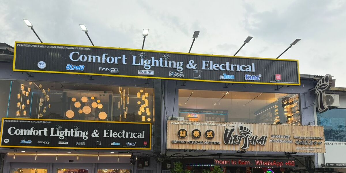Double shopfront with black signboards reading Comfort Lighting & Electrical and Pemborong Lampu dan Barangan Elektrik, brand logos below, and pendant lights glowing in the upper windows